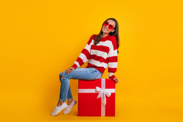 Full length photo of an excited beautiful stylish brunette woman in sunglasses while she is sitting and posing with a big red gift box