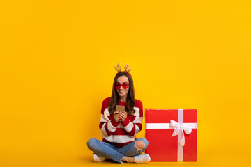 Full length photo of happy young smiling beautiful and charming woman in Christmas reindeer antlers while she sitting with big red gift box and using smart phone