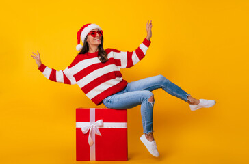 Full length photo of excited young smiling beautiful and charming woman in Santa hat and with Christmas mood while she sitting on big red gift box and having fun