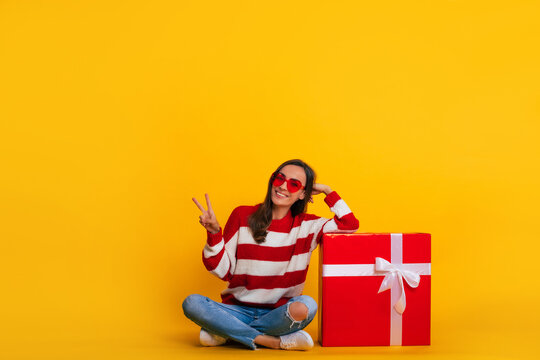 Full length photo of an excited beautiful stylish brunette woman in sunglasses while she is sitting and posing with a big red gift box