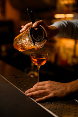hand of bartender gently holds a mixing cup with a strainer and pours liquid into wine glass