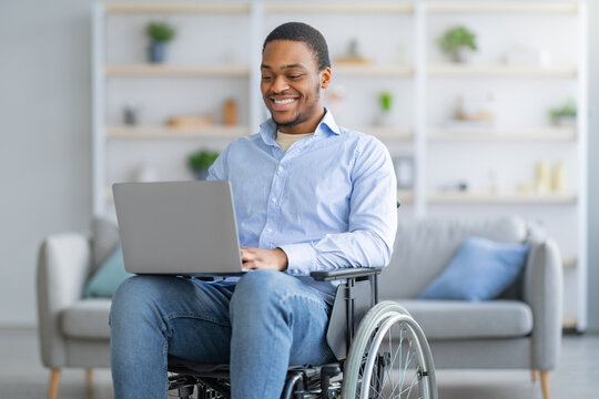 Portrait of cheerful disabled young black man in wheelchair using laptop for remote job at home