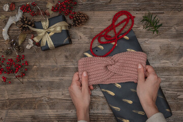 hands holding a handmade crochet headband as a christmas gift on a wooden surface with wrapping paper, ribbon, pine cones, berries and lights