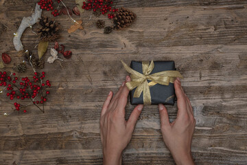 Hands holding a christmas gift on a wooden surface decorated with pine cones, berries and led lights