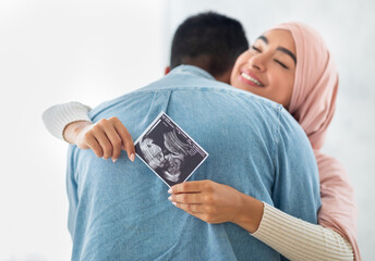 Expecting a baby. Loving pregnant arab woman holding ultrasound picture, hugging her husband and...