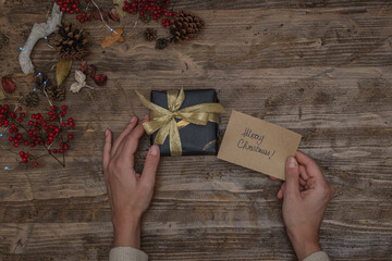 Flat lay of a christmas gift and female hands holding a greeting card on a wooden surface with pine cones, red berries and led lights
