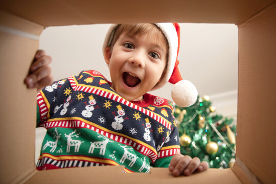Happy surprised Caucasian little boy in Santa's hat looking inside a cardboard box, at home near Christmas tree, bottom view. Excited little kid opening Christmas present.