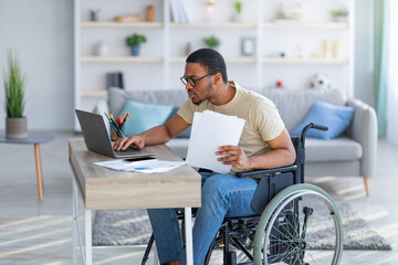 Paraplegic black guy in wheelchair with documents using laptop to work online from home