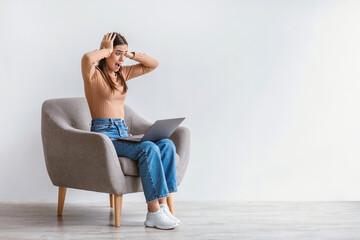 Portrait of shocked Caucasian woman sitting in armchair with laptop and grabbing head against white studio wall