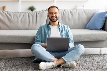 Full length of happy young guy sitting cross legged on floor, using laptop computer, smiling at camera indoors