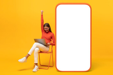 Winner. Young woman sitting on chair with her laptop, with huge phone mock up with blank screen