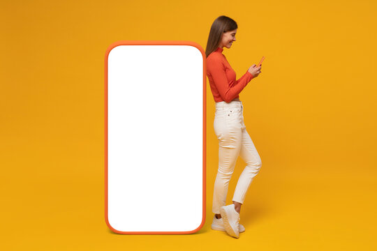 Young woman standing near huge phone mock up for app, isolated on yellow background