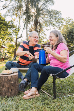 Smiling elderly spouses sharing smartphone and having picnic in nature