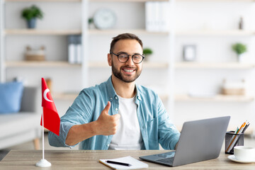 Happy young guy sitting at desk with flag of Turkey, using laptop, showing thumb up gesture at home