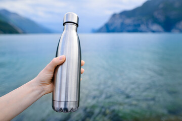 Close-up of female hand holding reusable, steel thermo shiny bottle for water, on the background of clear water of a lake with a turquoise hue. Copy space concept.