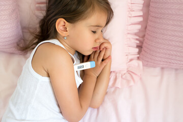 A sick child is lying in bed with an electronic thermometer under his arm with a high temperature. Cute small sleeps in a bed with pink pillows. Bed rest in case of illness. Fever and chills