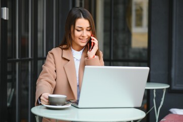 Young happy business lady looks at smartphone screen with toothy smile while sitting outside on terrace with laptop and coffee, working remotely online checking reports and getting ready for meeting