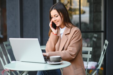 Young pretty business woman working on laptop in street cafe, typing on keyboard, smart lady seriously looking on screen.Smartphone and glasses on table. Wearing stylish pink jacket, white watches.