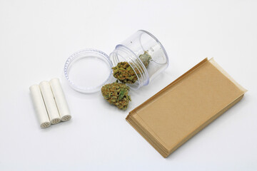 Marijuana buds in a clear pot accompanied by smoke paper and insulated filters on a white background
