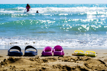 Three pairs of flip flops of different color, shape and size on a sandy beach on a sunny summer day with blurred blue sea and two unrecognizable bathing women in the background. Summer concept.