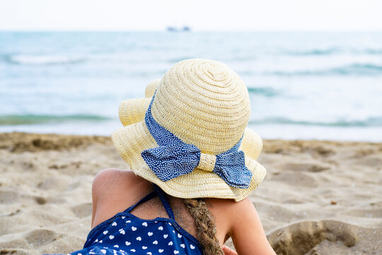 Close up from the back on a little girl wearing a straw hat with a blue polka dot bow, lying on a sandy beach, and looking at the sea with a blurred ship on the horizon.