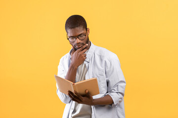 Thoughtful african american man in glasses reading book and touching chin, standing over yellow background