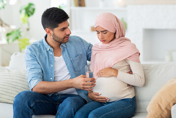 Care and support during pregnancy. Muslim expecting woman feeling sick, man giving her glass of...