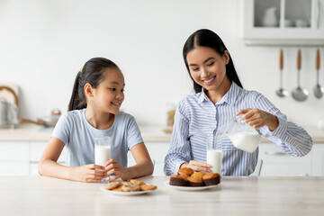 Young asian woman pouring calcium drink in glasses and having a bite together with daughter in kitchen