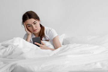 Sad european young woman suffering from depression and read message on smartphone sit on white bed at home
