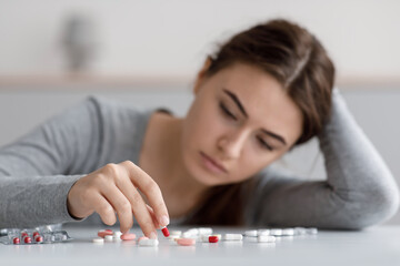 Indifferent caucasian young female suffering from depression, touching many scattered pills on table
