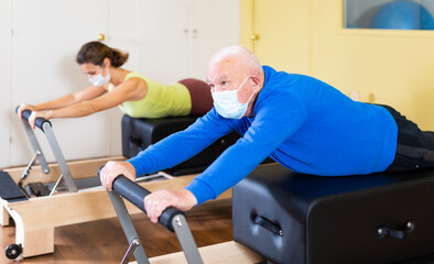 Mature man in protective mask practicing Pilates system on reformer supervised in gym