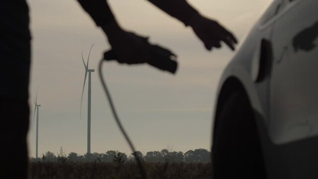 Wind driven generators charge ev on cloudy day. Man removes plug off white electric car against rotating turbines in field close view
