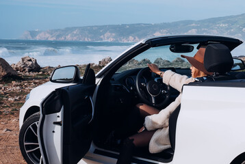 Outdoors lifestyle fashion portrait of stunning young woman driving cabriolet. Stop by the ocean. Girl travelling behind the wheel. Wearing stylish fur coat, sunglasses, hat. Travel by car. 