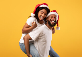 Happy black guy giving his girlfriend piggyback ride, wearing together Santa hats, having fun over yellow background