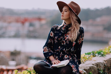 Outdoors lifestyle fashion portrait of pretty young woman sitting on old city wall. Reading book,...