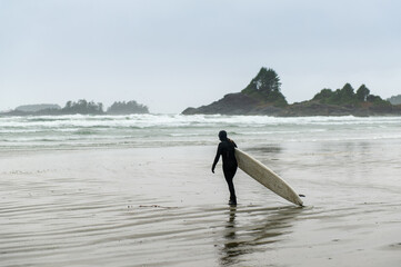 Anonymous surfer with surfboard walking on the beach of Cox Bay, a popular surf spot in Tofino...