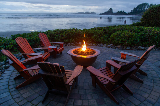 Adirondack chairs around a fireplace facing the beach of Cox Bay in Tofino Vancouver Island Canada. People are walking on the beach