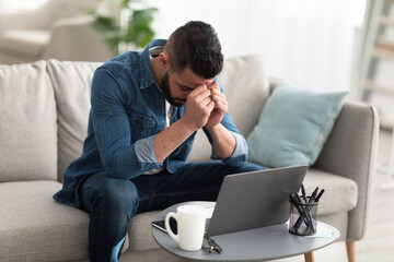 Fototapeta premium Stressed young Arab man sitting on couch near laptop, having problem with online work or studies at home