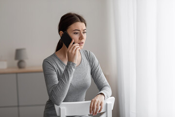 Sad unhappy worried european millennial female sitting on chair and talking on phone at home