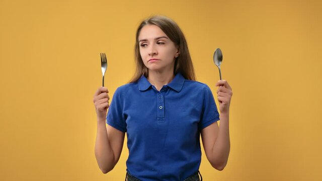 Portrait sad woman on diet choosing between fork and spoon suffering hungry isolated on orange 