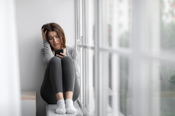 Despaired upset millennial millennial woman sitting on windowsill at home and reading message on phone, cry alone