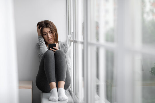 Despaired upset millennial millennial woman sitting on windowsill at home and reading message on phone, cry alone