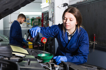 smiling young female is standing near car and replacing the oil in workshop.