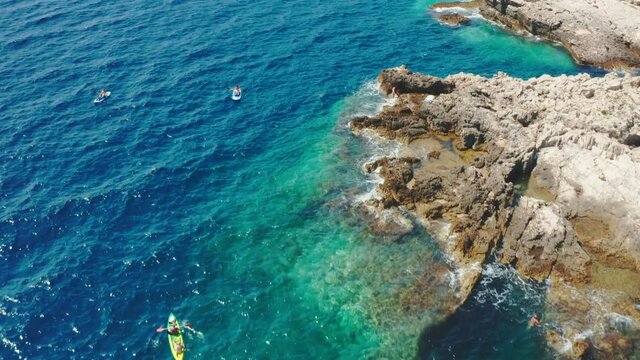 Sportsmen sail bright kayak and people on SUP surfboards in tranquil sea lagoon near limestone rocky beach on sunny day aerial view