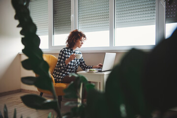 Caucasian woman in eyewear working on laptop while sitting on armchair near window holding a cup....