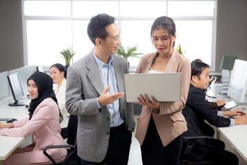 Asian businessman and businesswoman talking and looking laptop computer in the office, man standing explaining about financial with woman together, entrepreneur and employee planning about business.