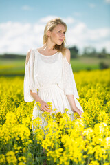 Naklejka na ściany i meble Happy girl posing in rape seed flowers field posing in white dress at sunny day outdoor