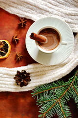 Winter warming drink. Cup of hot cocoa liquid chocolate with foam wrapped in white scarf. Top view on brown table decorated with cinnamon, star anise, dehydrated slice of lemon, fir cones and branch