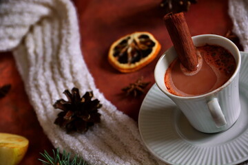 Winter warming drink. Cup of hot cocoa liquid chocolate with cinnamon stick close up. Brown table decorated with star anise, dehydrated slice of lemon, fir cones and branch, white scarf. Tilt horizon