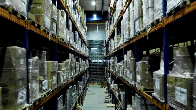 Drone flies between rows of stacked shelves in industrial warehouse, dolly out